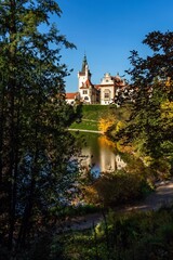 Pruhonice, Czech Republic - October 25 2020: View of the romantic castle surrounded with green and yellow trees standing in park. Reflection of the castle in water. Bright sunny autumn day, blue sky.