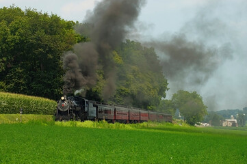 Fototapeta na wymiar Antique Steam Passenger Train Puffing Lots of Black Smoke along Amish Countryside with Green Fields