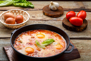 israeli fried eggs shakshuka with cheese in a frying pan next to products on the table.