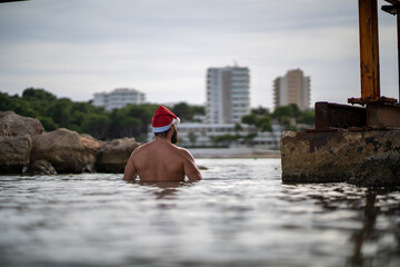 Fototapeta na wymiar A ma witth a Santa Claus hat stills standing between two rocks in the beach at Palmanova, with some hotels at the background during a winter day