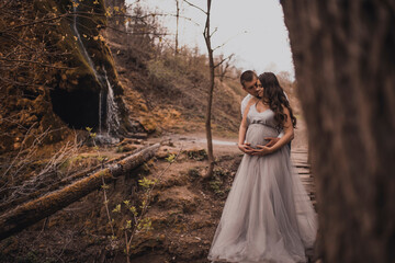 Fototapeta premium Family couple a man with a pregnant woman with a big belly in nature on the background of a waterfall. wooden bridge