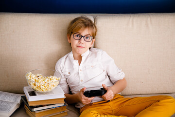 Blonde schoolboy boy plays video games, holds a gamepad, eats popcorn instead of learning lessons...