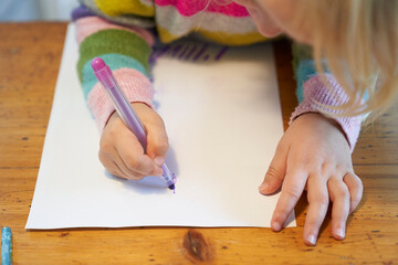 4 year old blonde girl with a brightly colored striped sweater sits at a wooden table and draws with a purple pencil on a piece of white paper