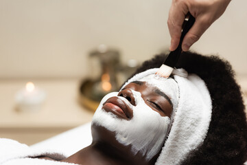African american woman in headband lying near spa therapist applying face mask on forehead on blurred background