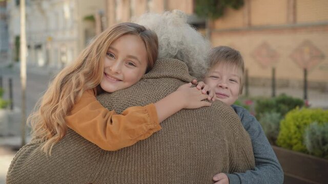 Happy children hugging male Caucasian retiree and smiling at camera. Portrait of joyful grandchildren meeting grandfather with long grey hair on sunny day outdoors. Childhood and retirement.