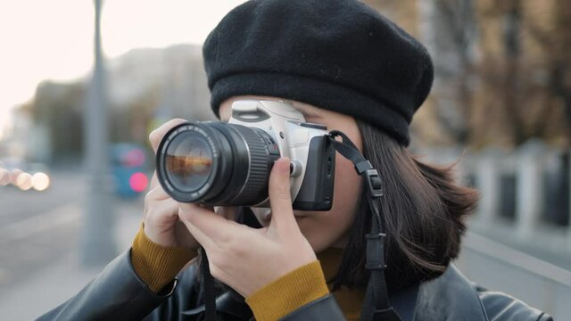 Young woman with photo camera doing pictures close up. Beautiful asian woman in black beret holding a camera shooting in the street, outside during an autumn day gimbal shot