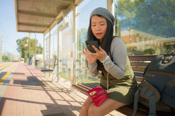 outdoors lifestyle portrait of young beautiful and happy Asian Japanese woman waiting the train sitting on station platform bench using mobile phone checking schedule online