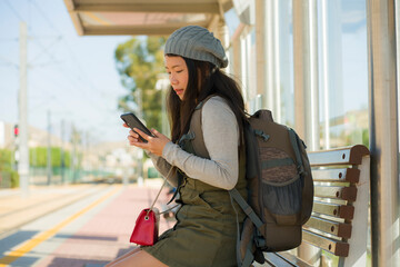 outdoors lifestyle portrait of young beautiful and happy Asian Korean woman waiting the train sitting on station platform bench using mobile phone checking schedule online