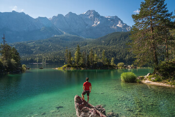 Boy in red shirt standing on a rock watching a beautiful view over lake Eibsee, in the Bavarian Alps. Famous touristic destination in the middle of the nature in Europe.