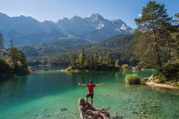 Boy in red shirt standing on a rock, spreading arms and watching a beautiful view over lake Eibsee, in the Bavarian Alps. Famous touristic destination in the middle of the nature.