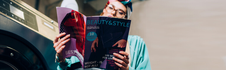 trendy woman in glasses and turban holding magazine in public laundromat, banner