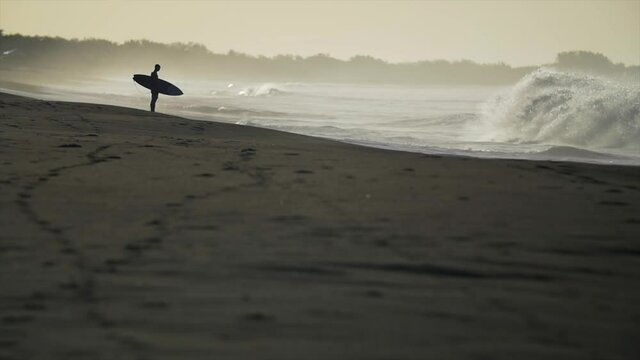 Silhouette of a surfer holding board with head down and kneeling, respecting power of ocean concept, large waves breaking on shore, epic surf lifestyle scene, slow motion, Nicaragua
