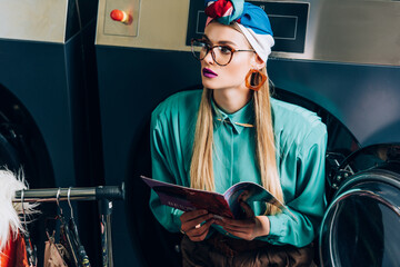 young woman in turban and glasses holding magazine in laundromat