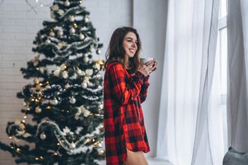 Pretty brunette girl standing near window, wearing red shirt and warm socks with cup of coffee and christmas lights behind.