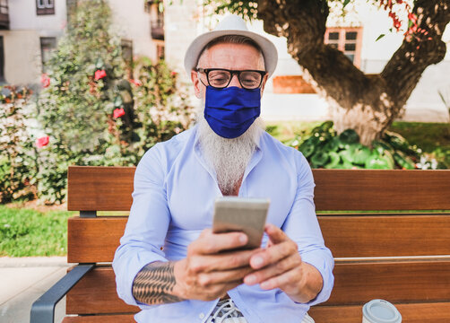 Cheerful senior hipster man with smartphone while wearing protective face mask for coronavirus - Outdoors