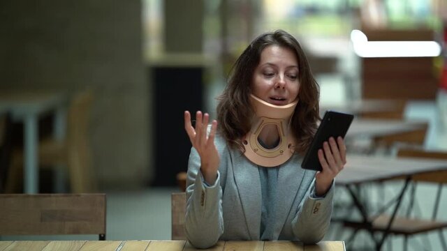 woman with neck brace is communicating online by video call on smartphone in cafe, curing of osteochondrosis