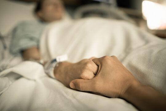 Person holding sick woman's hand lying in hospital bed.