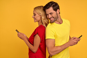 Side view of excited young couple two friends guy girl in red yellow t-shirts isolated on yellow background. People lifestyle concept. Standing back to back, using mobile phones, typing sms messages