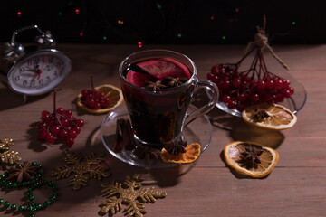 A Cup of fragrant mulled wine with spices, decorations for the New year, branches of red berries and a clock showing twelve o'clock, dark background. Horizontal orientation.
