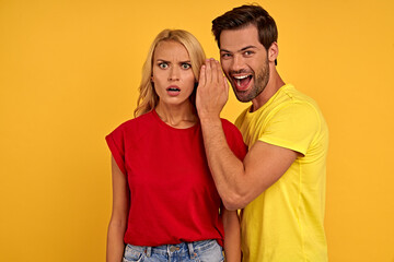 Naklejka premium Excited young couple friends guy girl in casual clothes isolated on yellow background. People lifestyle concept. Mock up copy space. Whispers gossip and tells secret behind hand, doing winner gesture