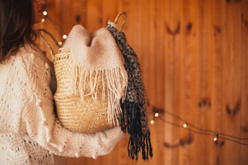 Woman in white sweater holding wicker basket with warm blanket in a wooden house with festive lights on the background.