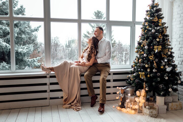 couple in love in festive clothers sitting and hugging near the large window and Christmas tree in decorated studio. happy holiday