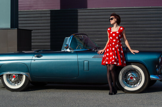 A young lady in a red polka dot dress and sunglasses stands near a vintage car.