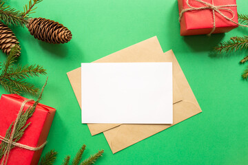 Christmas blank greeting card mock-up scene. Festive winter composition. Craft envelope, pine cone, gift box and fir tree branch on green background. Flat lay, top view, copy space.