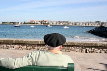 Persona anciana con txapela mirando hacia el mar. Sentado  en un banco con una pose natural