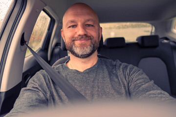 Bald male driver in focus, steering wheel out of focus. Man wears grey shirt, unshaven with grey and black beard in his 40s. Toned. Smiling expression on his face