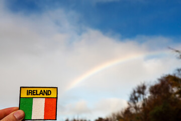 Badge with sign Ireland and National flag in focus, Blue cloudy sky with rainbow in the background. Irish luck concept.