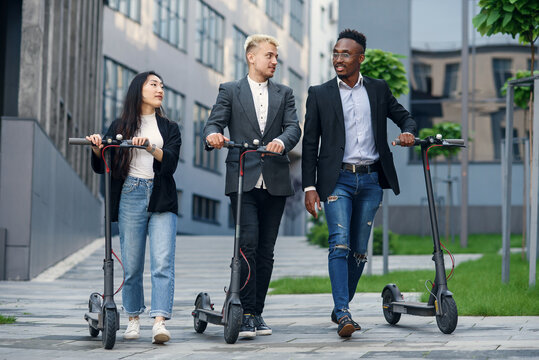 Confident happy multiracial office colleagues discussing business project go near office building with electric scooters