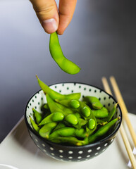 Hand picking up an edamame bean from a bowl. Fresh green soybeans in a bowl with chopsticks.
