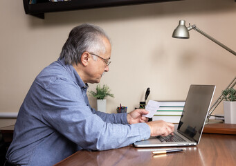 Senior man learn to use computer. Old man in glass and blue shirt Enters a password from a piece of...