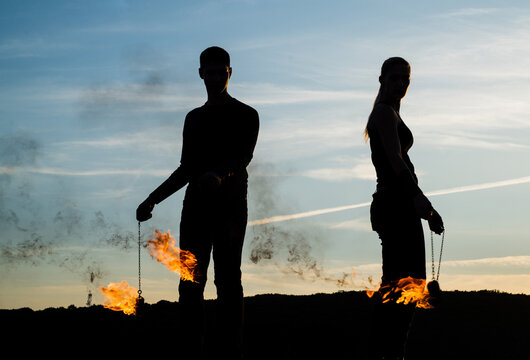 Couple of man and woman in dark silhouettes perform burning poi dance during fire performance on idyllic evening sky outdoors, smoke