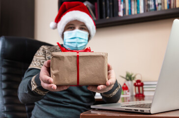 Christmas online congratulation. Young man in santa claus hat and face mask gives a gift and talks using laptop for video call friends and childrens. Christmas during coronavirus.