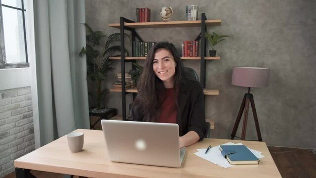 Young woman employee works in the office at the computer smiling. Brunette girl looking at laptop screen, working remotely at home. Work during self-isolation and quarantine COVID-19 4k