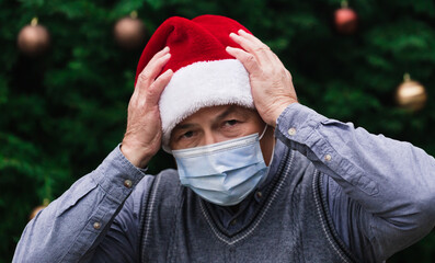 Shocked or surprised Christmas. Close up Portrait of senior man wearing a santa claus hat and medical mask with emotion. Against the background of a Christmas tree. Coronavirus pandemic