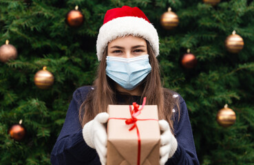 Christmas mask congratulations. Portrait woman wearing santa hat and blue sweater in medical mask, giving gift present box with red ribbon, christmas tree bokeh on background