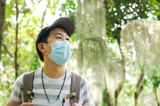 Young adult Asian travel backpacker wearing a face mask hiking in the forest. Millenial man wearing a cap walking in the nature during Covid-19 or Corona Virus pandemic.