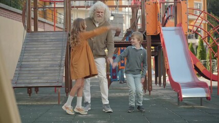 Wide shot of positive senior man. boy and girl stacking hands and start playing hide-and-seek. Grandfather closing eyes as children running away to hide. Happy weekend family leisure.
