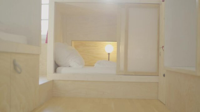 Shot of a bed in a beautiful wooden modern youth hostel with a warm lamp shining on white sheets. Smooth backwards camera dolly movement.