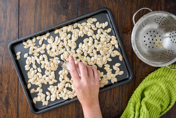 Preparing to roast fresh pumpkin seeds, woman’s hand salting washed seeds on a baking sheet 
