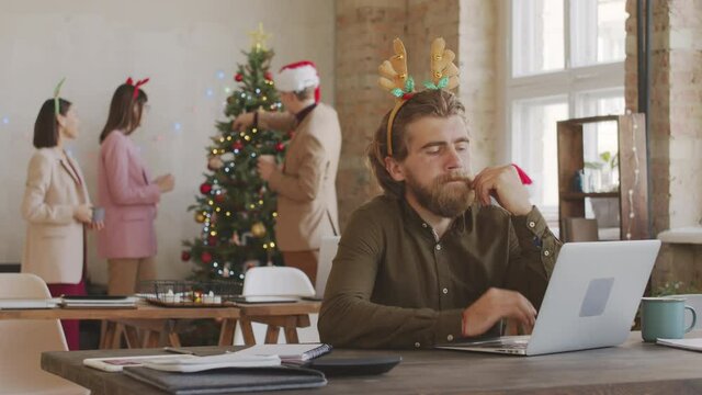 Medium shot of stressed man in festive headband working on laptop in office on Christmas. He is rubbing his eyes in exhaustion and looking at screen as his happy colleagues chatting by Christmas tree