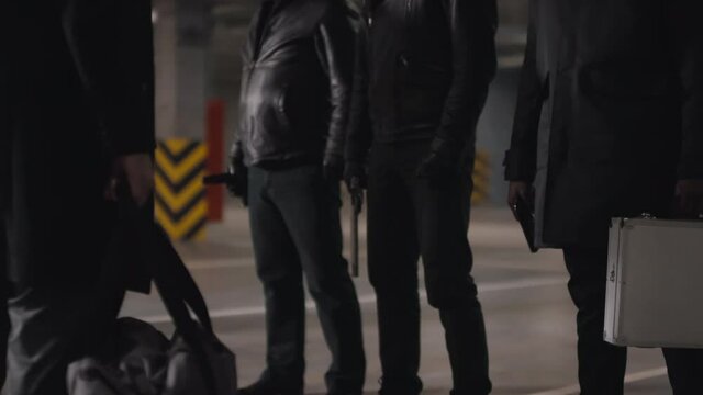 Sequence of shots of assertive multi-ethnic bandits meeting at dark underground parking transmitting briefcase with money