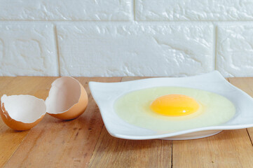 Eggs hammered on a white plate with an egg shell on the side, placed on a wooden table in the kitchen with a brick background.
