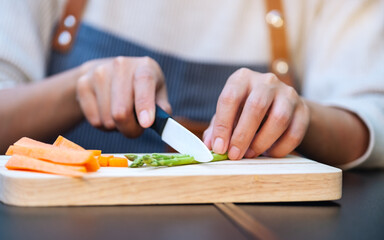 Closeup image of a woman cutting and chopping asparagus and carrot by knife on wooden board