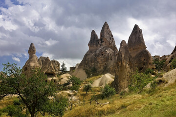 Fototapeta premium Against the background of a cloudy sky, sharp-pointed rocks with ancient caves. Around the yellowed grass, trees. Cappadocia in summer. Turkey.