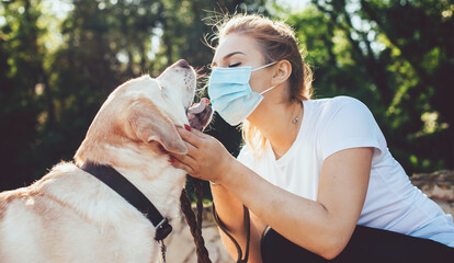 Blonde woman with medical mask on face is playing with her golden retriever in a park during a summer walk