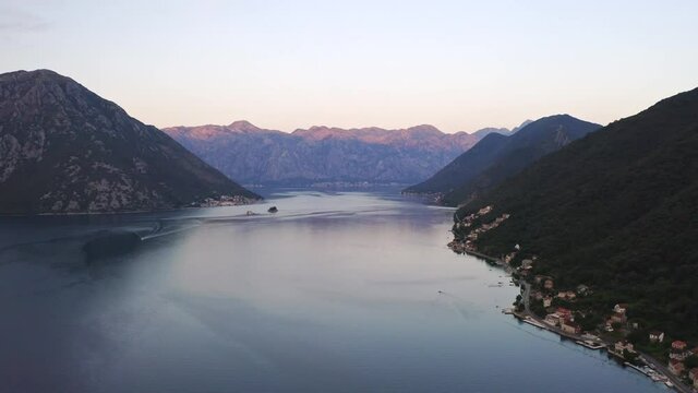 The bay of Kotor in Montenegro with a coastal town on its shore, mountain ranges surrounding the bay and dominating the horizon, cloudless sky above, red morning glow shining on its peaks, aerial.